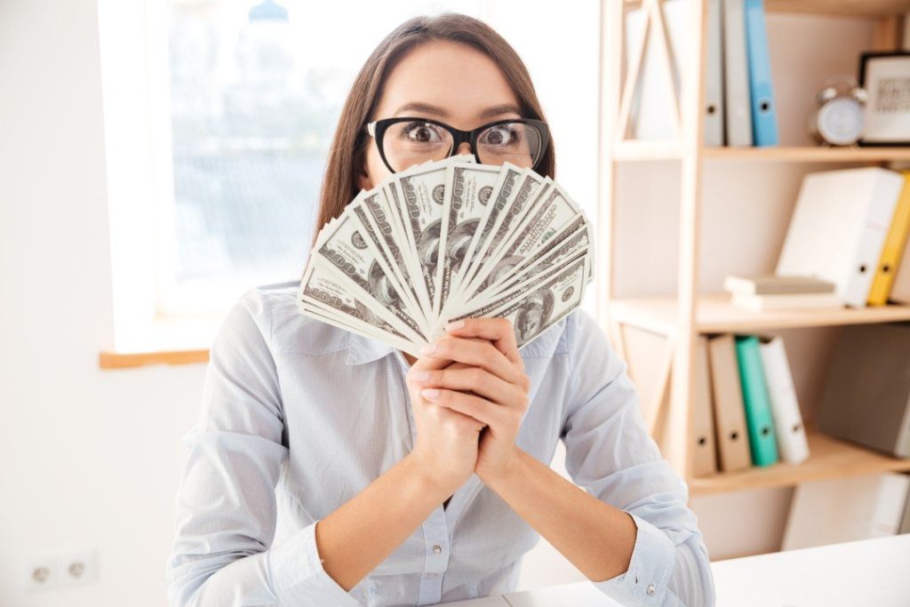 A businesswoman in an office holding a fan of one hundred dollar bills up to her face