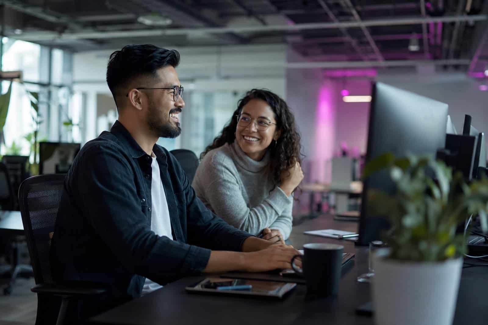 Two colleagues smiling together at a workstation in a modern office, signalling a settled, lasting hire